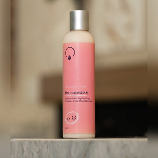 A bottle of The Condish System Free Conditioner with a pink label and silver cap, standing on a marble surface with a blurred indoor background.