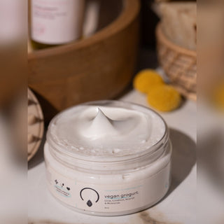 A white jar of vegan arogurt body butter with a swirl of cream visible on top, set on a marble surface with a wooden tray and yellow sponge in the background.